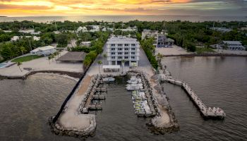 An aerial view of a waterfront property with a centrally located building, boat docks, surrounding greenery, and a stunning sunset in the background.