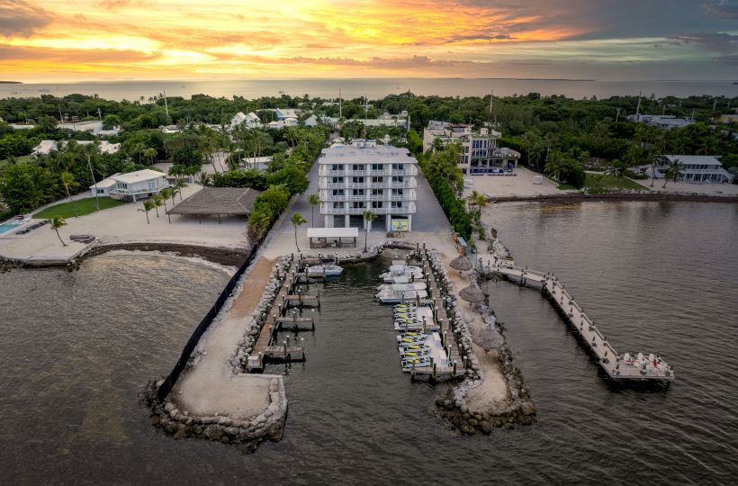 An aerial view of a waterfront property with a centrally located building, boat docks, surrounding greenery, and a stunning sunset in the background.