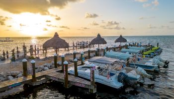 Boats are docked at a marina with thatched-roof huts and a scenic sunset in the background. The calm water reflects the golden hues in the sky.