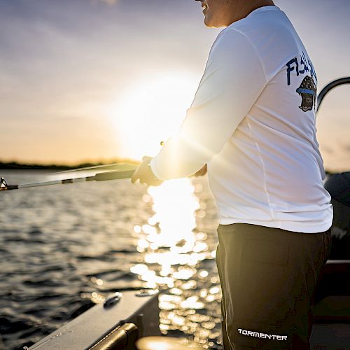 A person is on a boat fishing at sunrise or sunset, dressed in a white long-sleeve shirt and dark shorts.