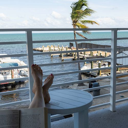 Someone is relaxing, reading a book on a balcony with a view of the ocean, boats, and a palm tree swaying in the breeze, feet propped up.
