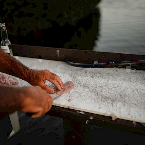 A person is preparing to slice fish on a cutting board outside near a body of water. A drink and a knife are visible on the counter.