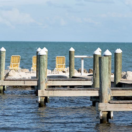 A wooden dock with posts extends into calm waters, with two yellow chairs and a small table visible on the shoreline in the distance.