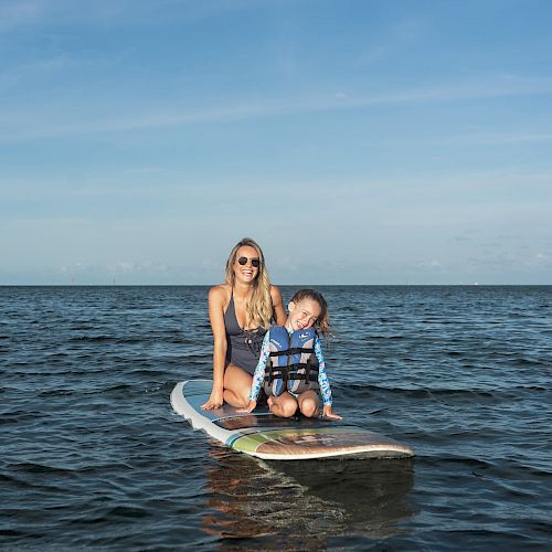 A woman and a child, both wearing swimwear, are sitting together on a paddleboard in calm waters under a clear sky.