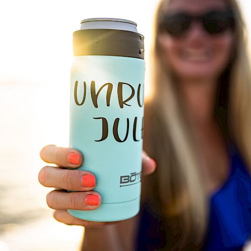 A person with sunglasses and long blonde hair holds a can wrapped in a light blue koozie with the words "UNRU JUL." The background is blurry.
