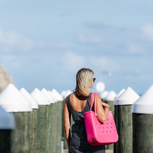 A person with blonde hair, wearing a striped dress, is walking on a pier, carrying a pink bag, with a thatched hut and blue sky visible in the background.