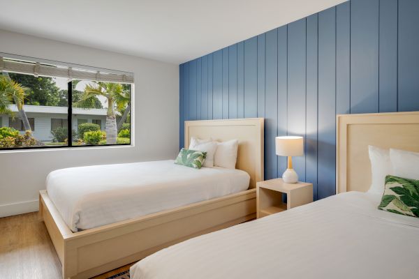 The image shows a bedroom with two beds, blue accent wall, bedside lamp, and a window overlooking greenery outside.