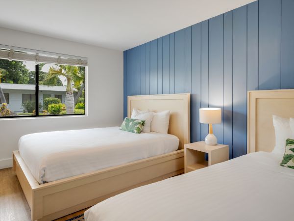 The image shows a bedroom with two beds, blue accent wall, bedside lamp, and a window overlooking greenery outside.