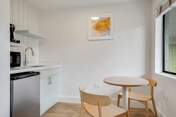 A small kitchen area with a sink, mini-fridge, microwave, coffee maker, and two wooden chairs around a round table. A fish painting is on the wall.