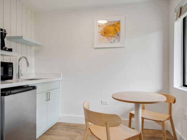 A small kitchen area with a sink, mini-fridge, microwave, coffee maker, and two wooden chairs around a round table. A fish painting is on the wall.
