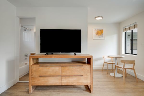 A room with a TV on a wooden stand, a small table with two chairs, a bathtub in the corner, and a window letting in natural light.