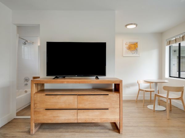 A room with a TV on a wooden stand, a small table with two chairs, a bathtub in the corner, and a window letting in natural light.