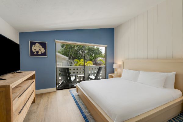 A modern bedroom with a large bed, wooden furniture, a TV, a blue accent wall, and a window leading to a balcony with outdoor seating.