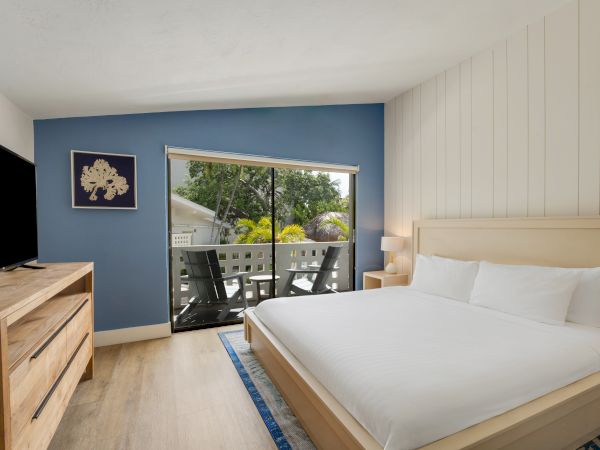 A modern bedroom with a large bed, wooden furniture, a TV, a blue accent wall, and a window leading to a balcony with outdoor seating.