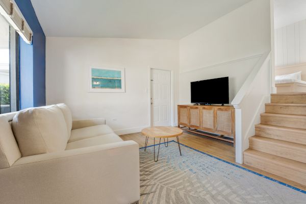 A modern living room features a beige couch, a wooden coffee table, a TV on a wooden stand, an area rug, and wooden stairs leading upward.