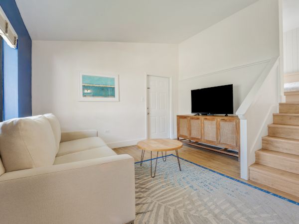 A modern living room features a beige couch, a wooden coffee table, a TV on a wooden stand, an area rug, and wooden stairs leading upward.