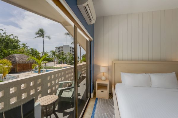 A cozy hotel room with a bed, nightstand, and balcony overlooking a palm tree-lined pool area. The large window offers a scenic outside view.