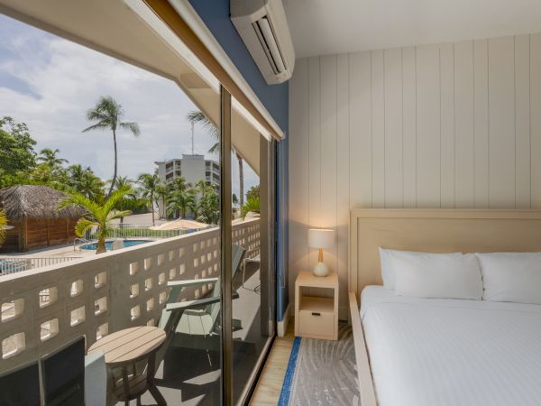 A cozy hotel room with a bed, nightstand, and balcony overlooking a palm tree-lined pool area. The large window offers a scenic outside view.