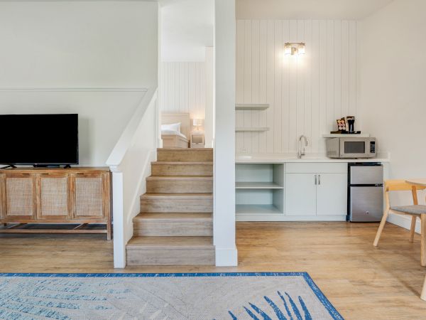 A modern interior featuring a split-level room with a TV, kitchenette, dining area, stairs leading to a bed, and a cozy carpet on wooden flooring.