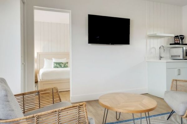 The image shows a simple, modern living area with wicker chairs, a round wooden table, a wall-mounted TV, kitchen appliances, and a view into a bedroom.