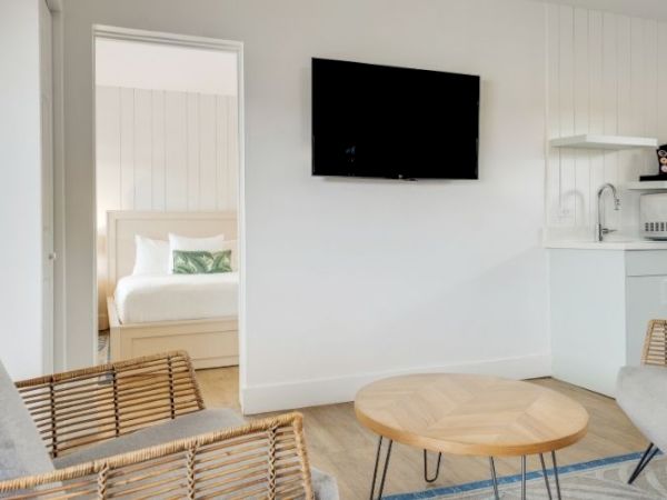 The image shows a simple, modern living area with wicker chairs, a round wooden table, a wall-mounted TV, kitchen appliances, and a view into a bedroom.
