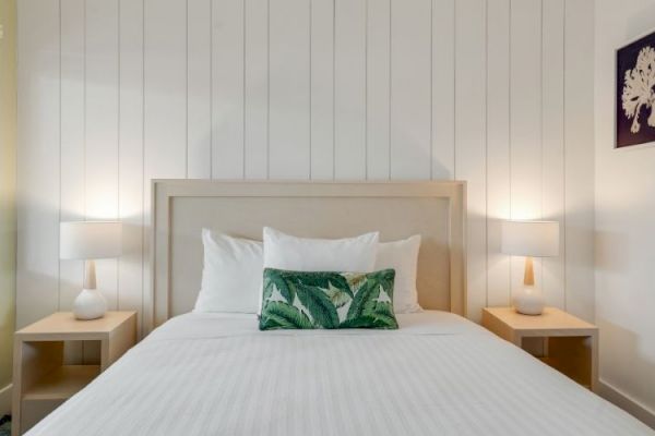 A cozy bedroom with a white-paneled wall, a double bed with a green-leaf pillow, and two bedside tables with lamps. A window is on the left.