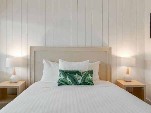 A cozy bedroom with a white-paneled wall, a double bed with a green-leaf pillow, and two bedside tables with lamps. A window is on the left.