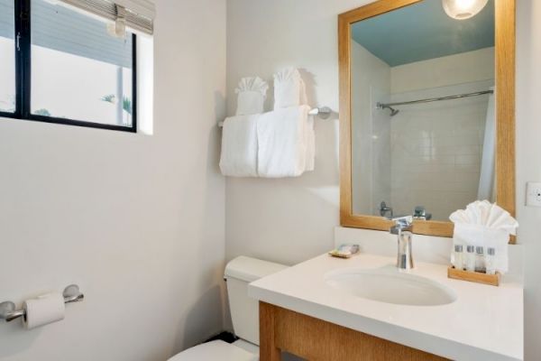 A clean bathroom with a sink, mirror, toilet, towel rack with folded towels, a shower curtain, a small window, and toiletries by the sink.