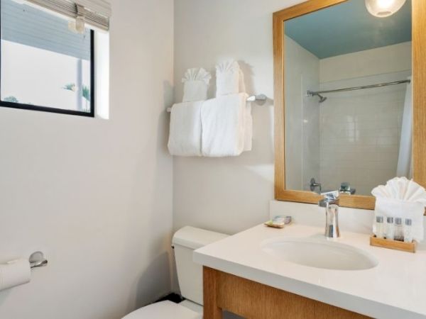 A clean bathroom with a sink, mirror, toilet, towel rack with folded towels, a shower curtain, a small window, and toiletries by the sink.
