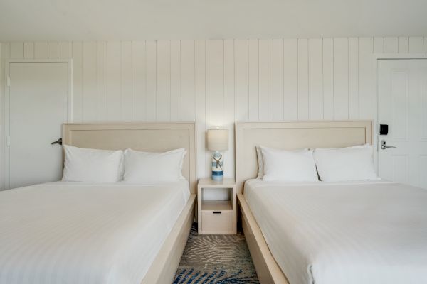 A hotel room with two twin beds, a nightstand with a lamp and phone, and white paneled walls is shown.