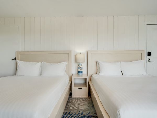A hotel room with two twin beds, a nightstand with a lamp and phone, and white paneled walls is shown.