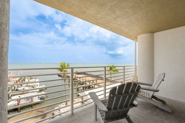 A balcony with two Adirondack chairs overlooks a marina and the ocean under a partly cloudy sky.
