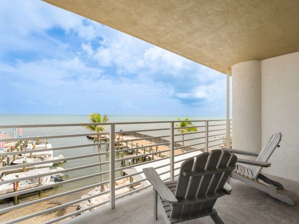 A balcony with two Adirondack chairs overlooks a marina and the ocean under a partly cloudy sky.