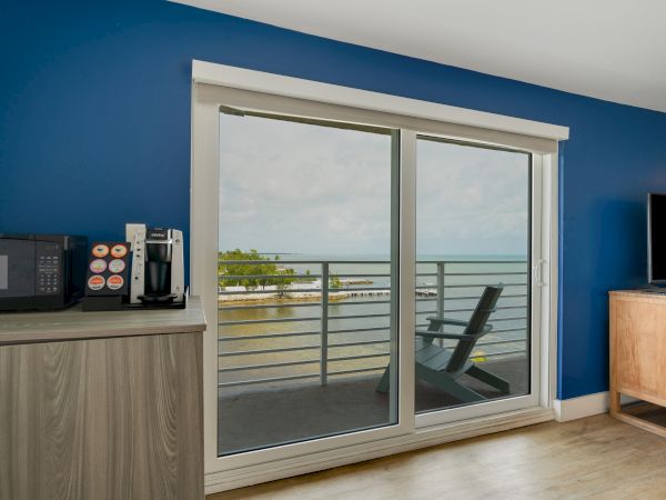 A room with a blue wall has a microwave, coffee machine, TV, and a sliding door leading to a balcony overlooking water with trees in the background.