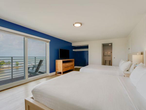 A hotel room with two beds, a TV, dresser, balcony with chairs, and an ocean view is shown. Walls are blue and white, decor is minimalist.
