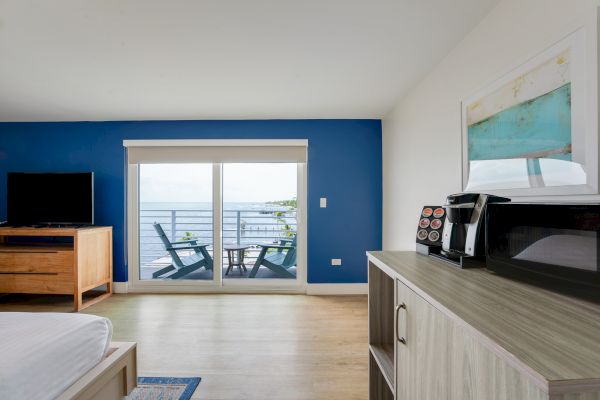 A bright room with a TV, microwave, coffee maker, two chairs on a balcony facing the ocean, and a blue accent wall.