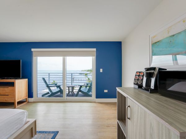 A bright room with a TV, microwave, coffee maker, two chairs on a balcony facing the ocean, and a blue accent wall.
