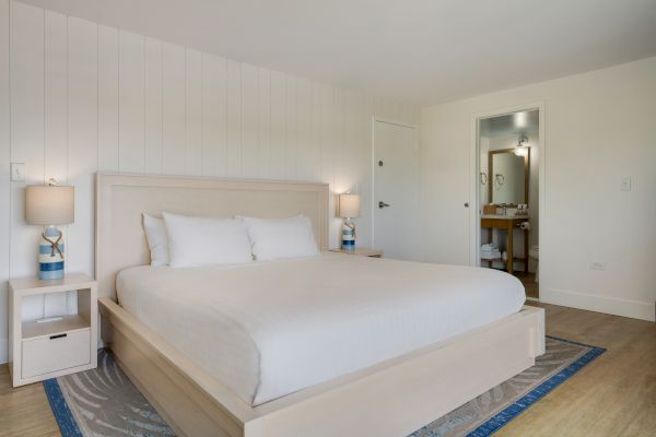 A modern bedroom features a white bed, two side tables with lamps, and a bathroom visible through an open door.