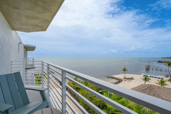 A coastal view from a balcony with two chairs. The scene includes a beach, a dock, palm trees, and a partly cloudy sky over the calm ocean.
