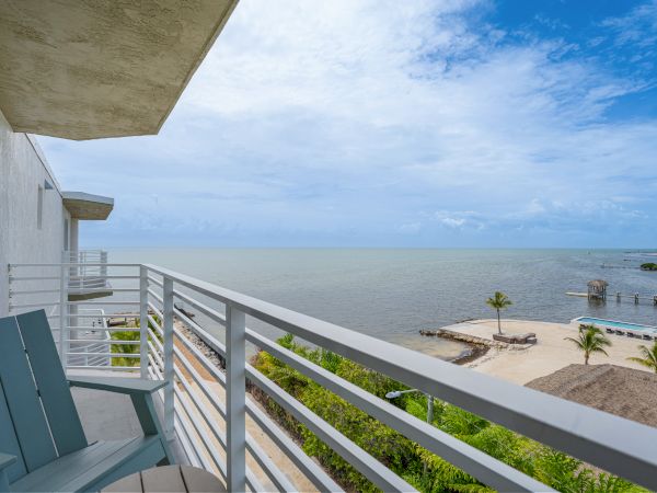A coastal view from a balcony with two chairs. The scene includes a beach, a dock, palm trees, and a partly cloudy sky over the calm ocean.