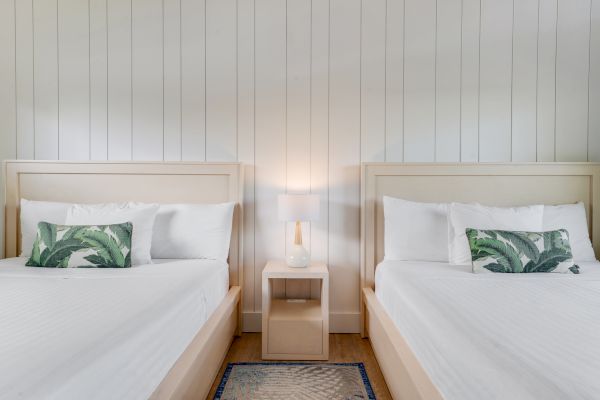 The image shows a clean, minimal bedroom with two white beds, green-leaf pillows, a small white nightstand, and a lamp in the center.