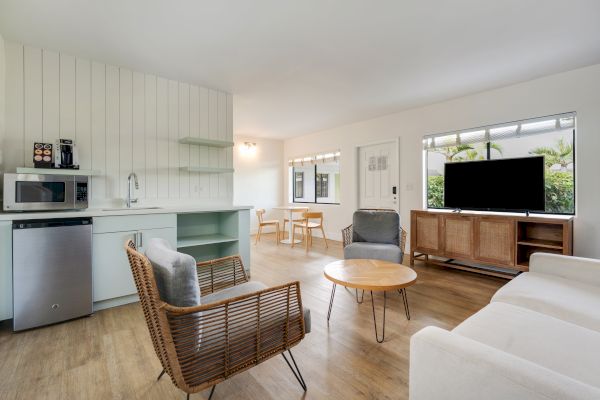 A modern living room with a TV, wicker chairs, a sofa, and a small kitchen area with a microwave and mini-fridge, leading to a dining space.