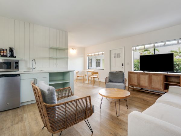 A modern living room with a TV, wicker chairs, a sofa, and a small kitchen area with a microwave and mini-fridge, leading to a dining space.