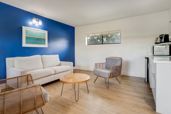 A modern living room with a white sofa, blue accent wall, wall art, wicker chairs, a wooden coffee table, and a kitchenette in the background.