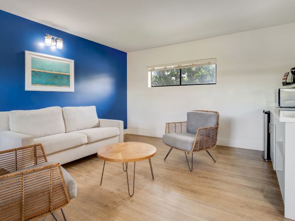 A modern living room with a white sofa, blue accent wall, wall art, wicker chairs, a wooden coffee table, and a kitchenette in the background.