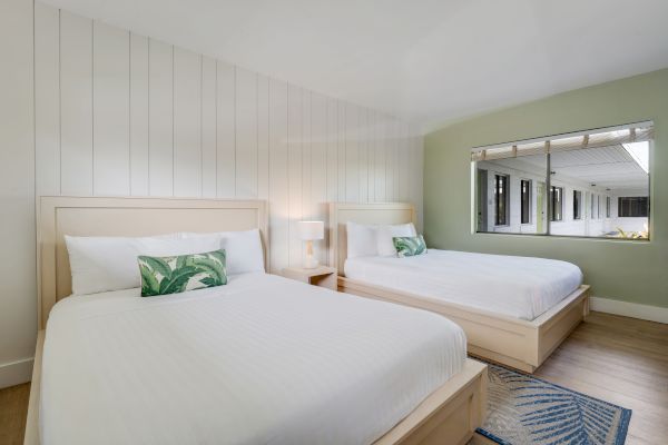 A modern hotel room features two large beds with white linens and decorative pillows, a nightstand with a lamp, and a window with an exterior view.