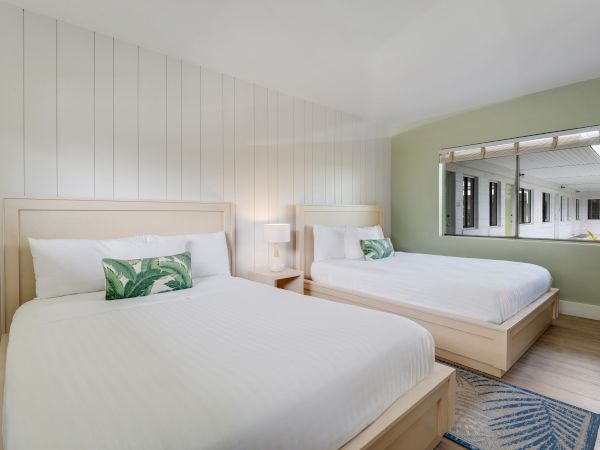 A modern hotel room features two large beds with white linens and decorative pillows, a nightstand with a lamp, and a window with an exterior view.