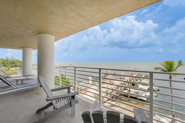 A balcony with chairs overlooks a marina and ocean under a cloudy blue sky. Palm trees and docks are visible near the water's edge.