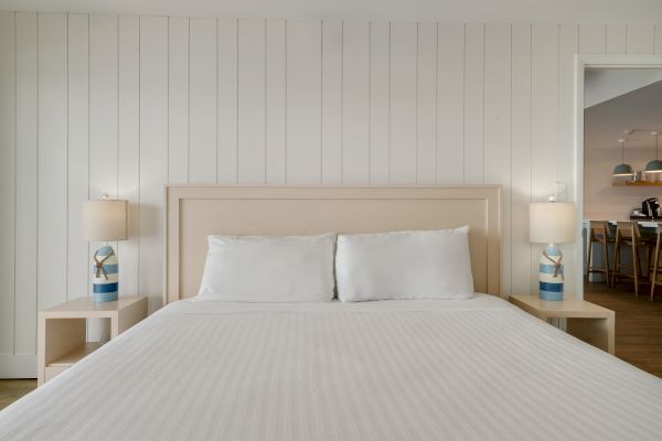 A neatly made bed with white linens is flanked by two lamps on wooden nightstands; a glimpse of a dining area is visible through an open doorway.