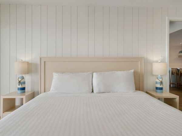 A neatly made bed with white linens is flanked by two lamps on wooden nightstands; a glimpse of a dining area is visible through an open doorway.